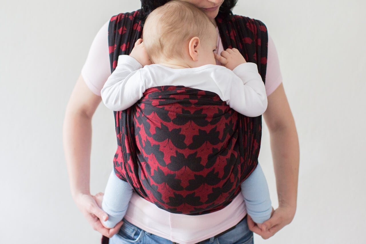 A cropped view of a baby in a woven wrap. A front view of a baby wearing mother carrying her small child in a sling.