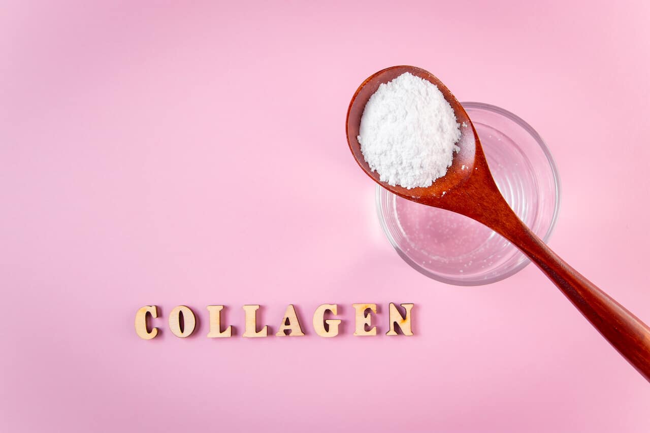 Natural bio supplement collagen powder in wooden spoon and glass of pure water on pink background. Concept healthy skin and bones, anti-aging extra protein product. Flatlay, place for text.