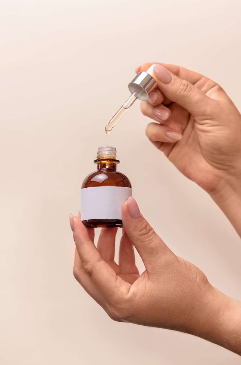 Hands holding pipette and opened dropper bottle with blank label over light beige, close up, mockup. Natural cosmetic, serum or essence. Everyday woman skincare routine