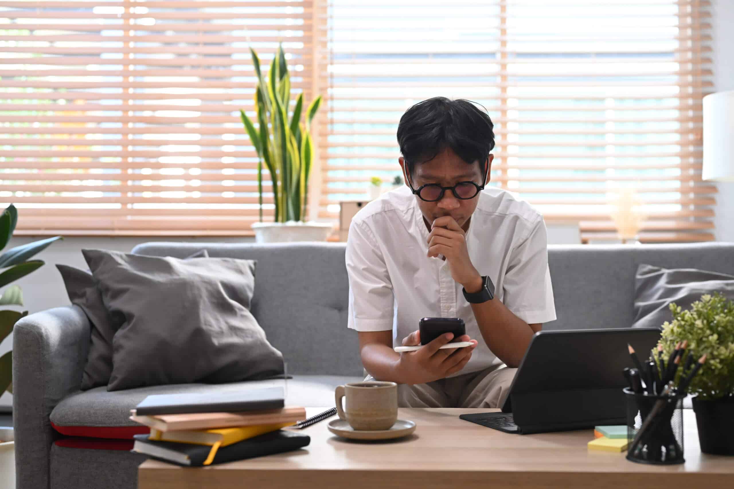 Man sitting on couch in living room and using smart phone for shopping online, checking social media.