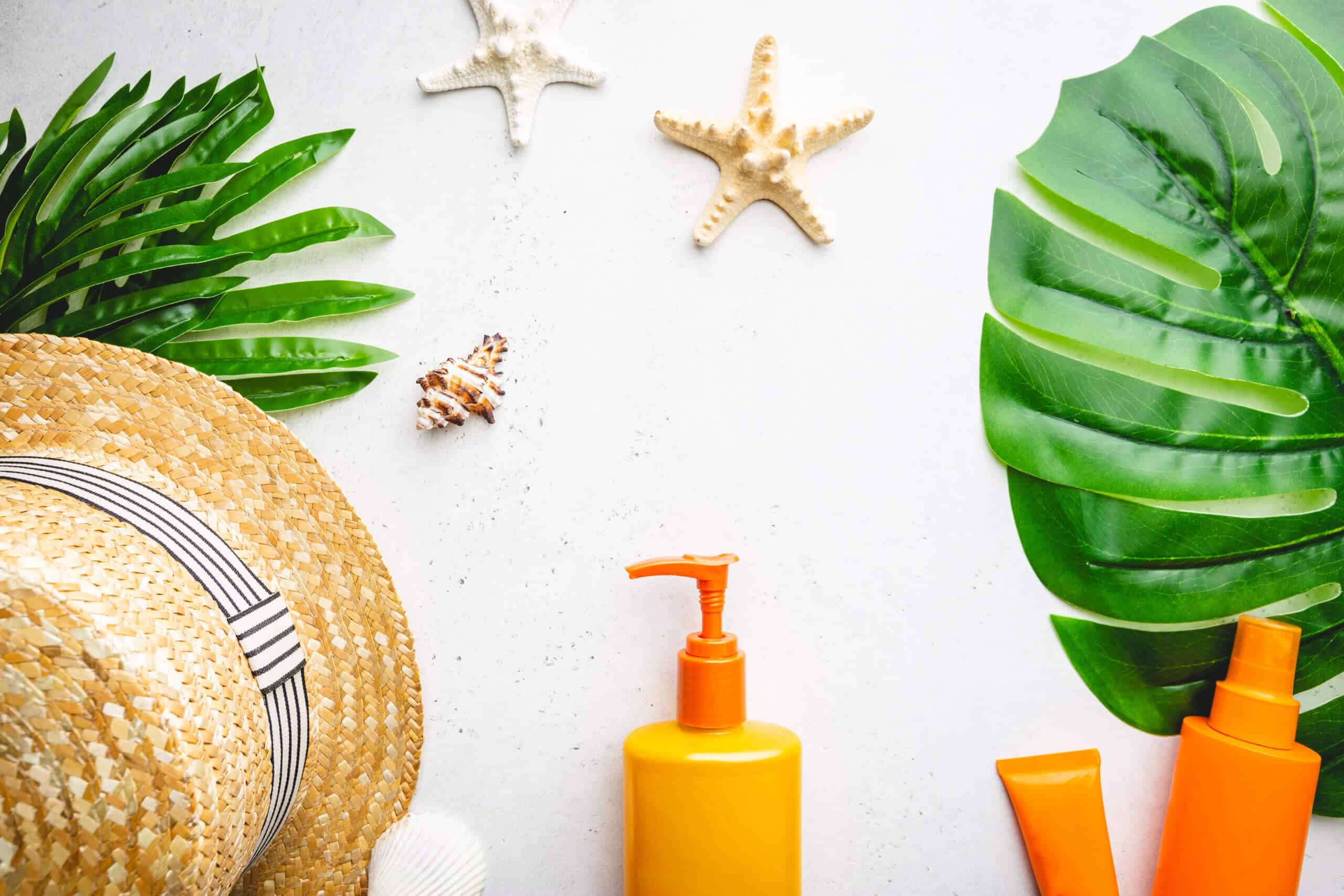 Sunscreen bottles arranged with straw hat on white background with palm leaves, top view