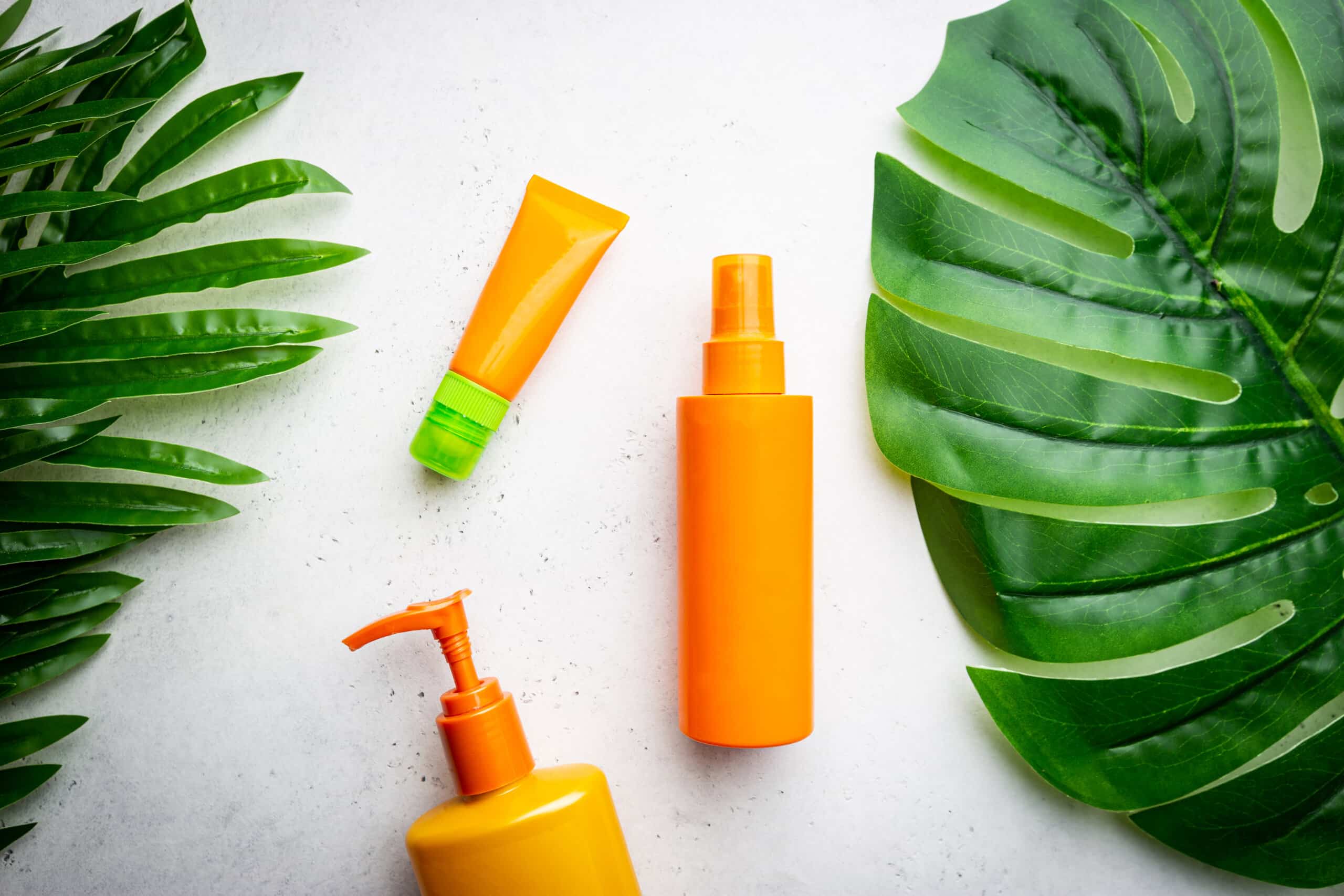 Sunscreen bottles arranged on white background with palm leaves, top view
