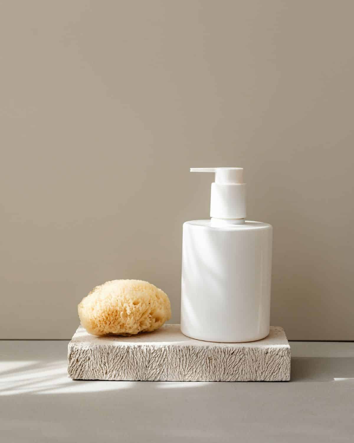 Bodylotion-Flasche neben einem Reinigungsschwamm auf einer neutralen Oberfläche.