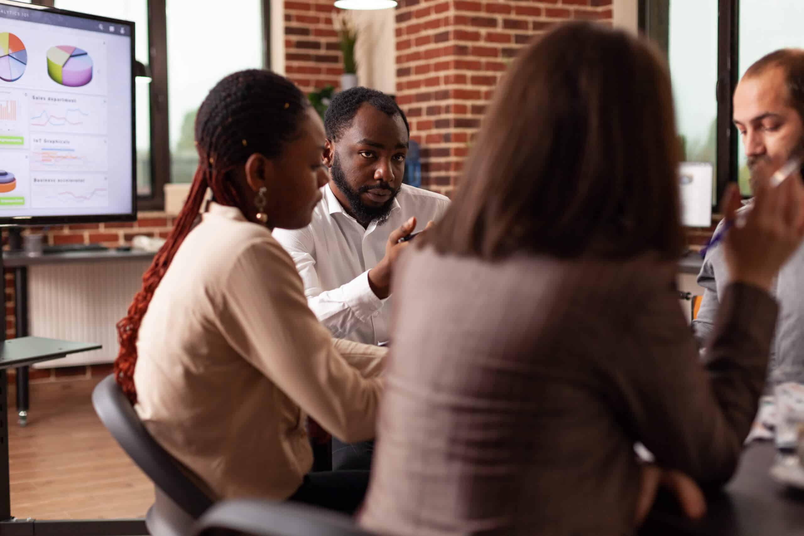 Afroamerikanischer Mann erklärt die Strategieplanung des Lieferantenmanagements während einer Geschäftspräsentation in einem Startup-Büro. Diverse Geschäftsleute arbeiten zusammen und brainstormen Ideen für eine Partnerschaft