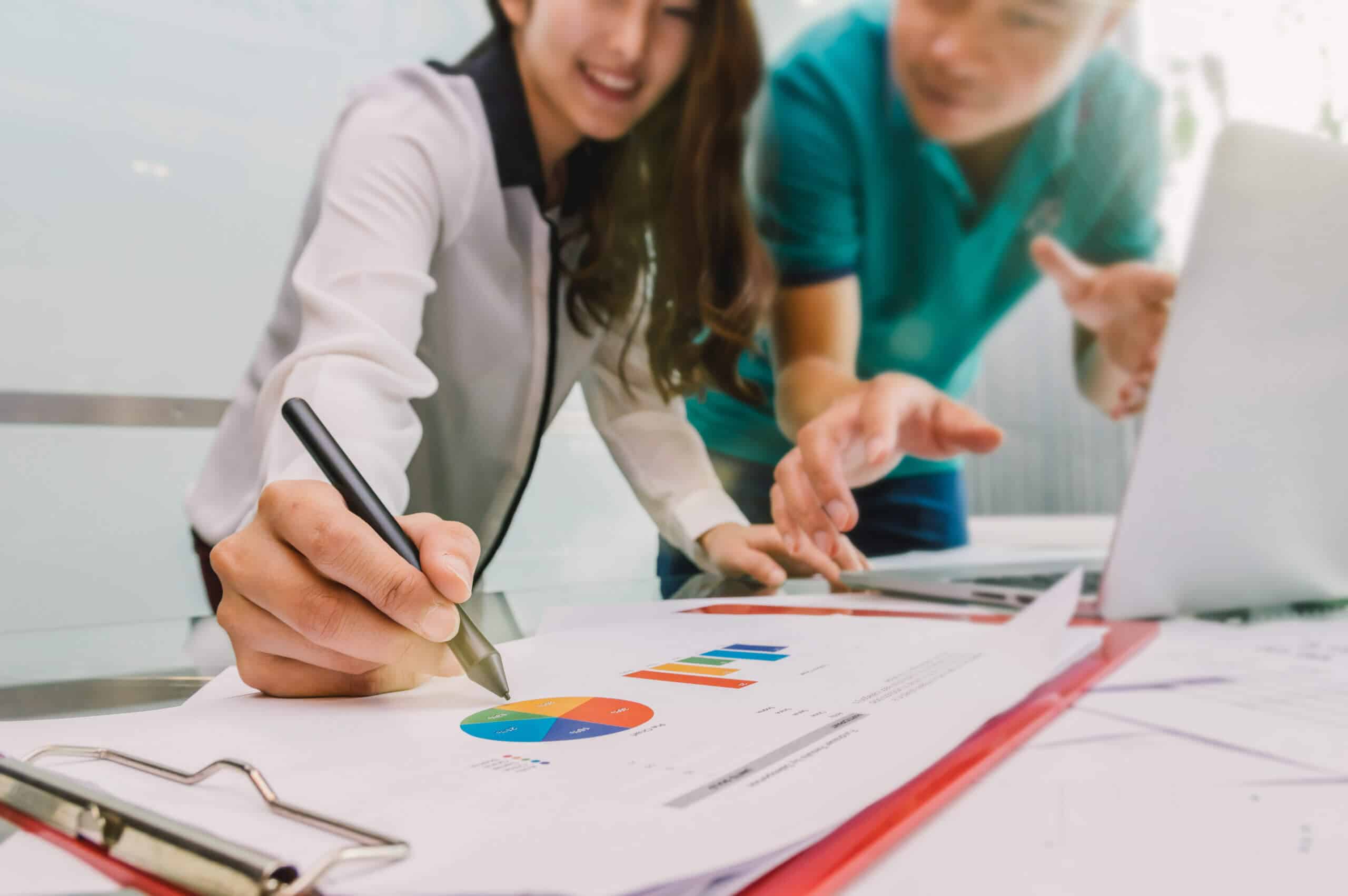 Geschäftsdokumente mit Laptop am Arbeitsplatz, im Hintergrund arbeitet ein Team im Besprechungsraum an der ISO 9001-Zertifizierung.