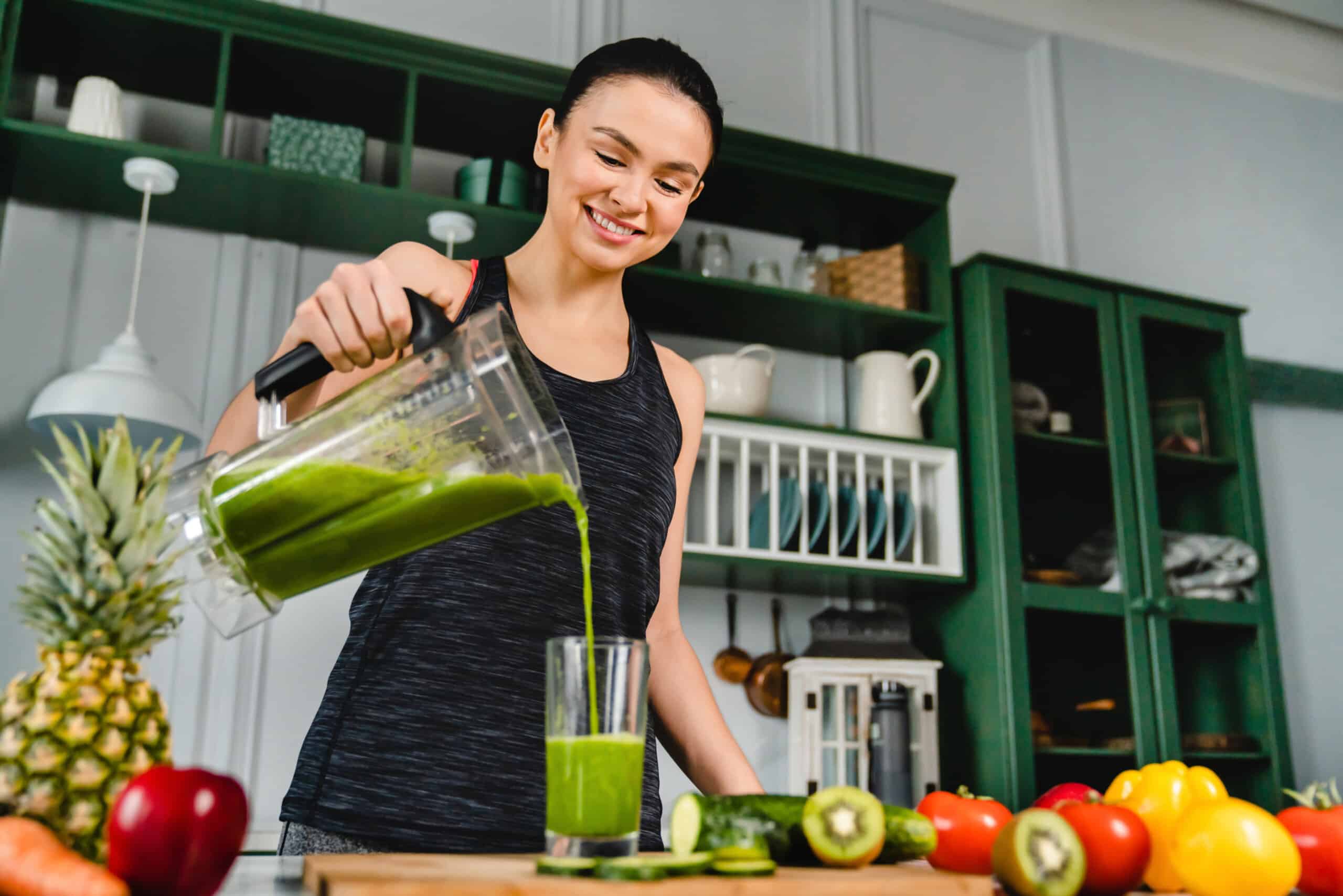 Glückliches, junges, schlankes Mädchen macht in der Küche mit einem Mixer einen frischen, biozertifizierten Smoothie