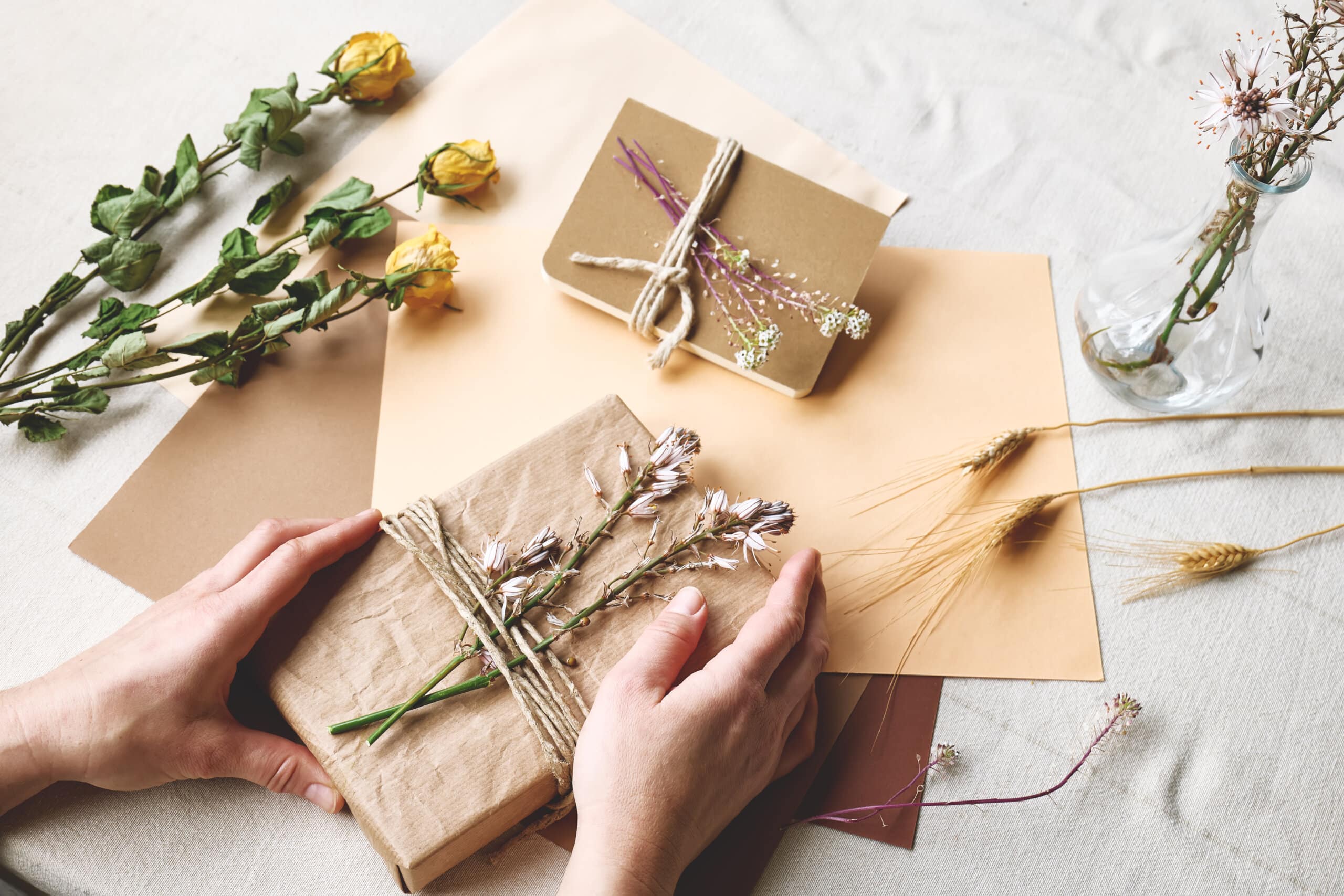 Frau stellt ein abfallfreies, plastikfreies, trendiges, handgemachtes Geschenkpaket mit recyceltem Bastelpapier und Trockenblumen auf dem Tisch mit Leinentischtuch her. Natürliche Ästhetik für biozertifizierte Produkte.