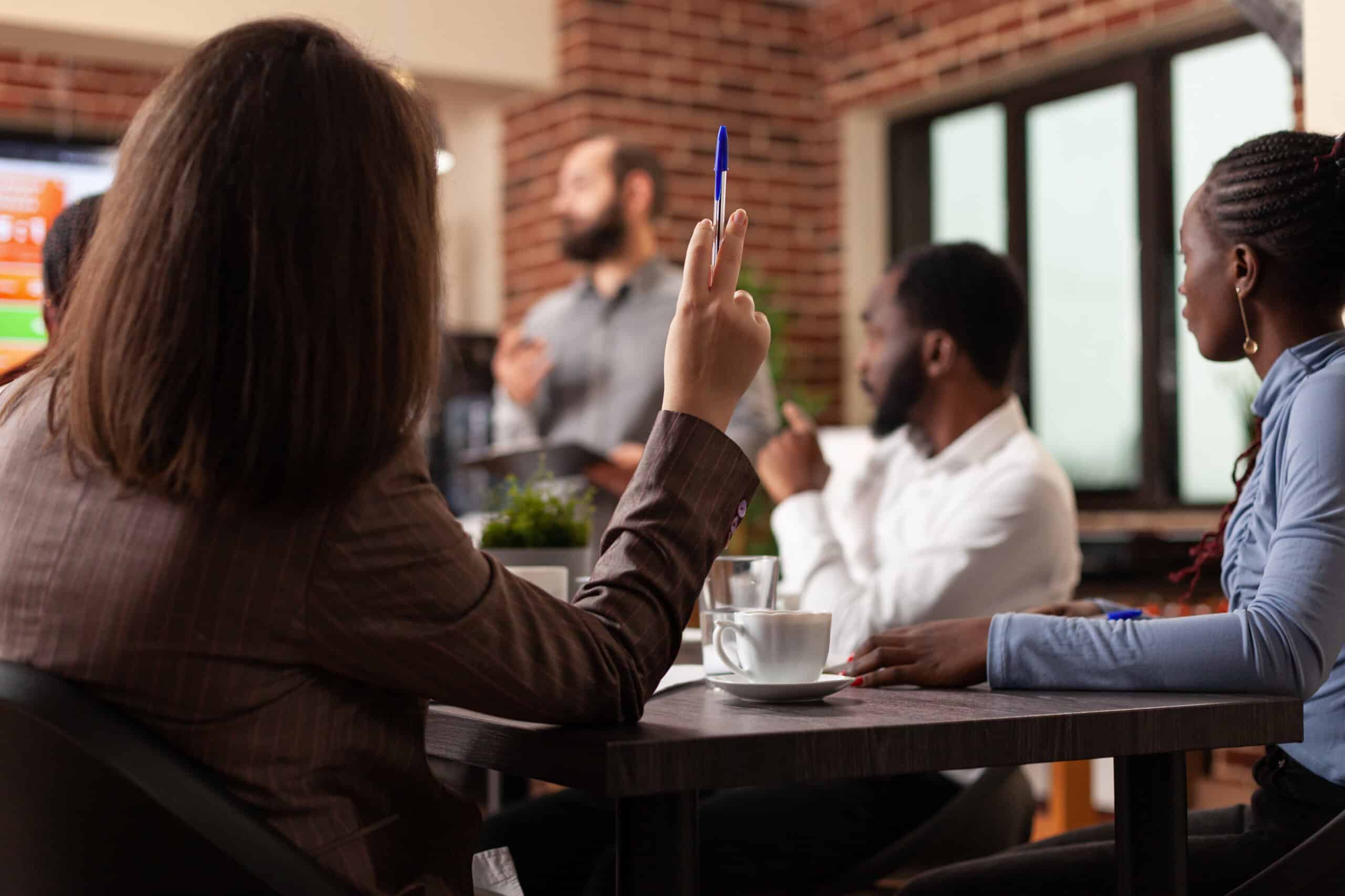 Diverse Geschäftsleute arbeiten an einer Managementpräsentation und analysieren Firmengrafiken in einem Startup-Büro. Geschäftsfrau hebt die Hand und hat eine Geschäftsfrage zu einem Partnerschaftsprojekt