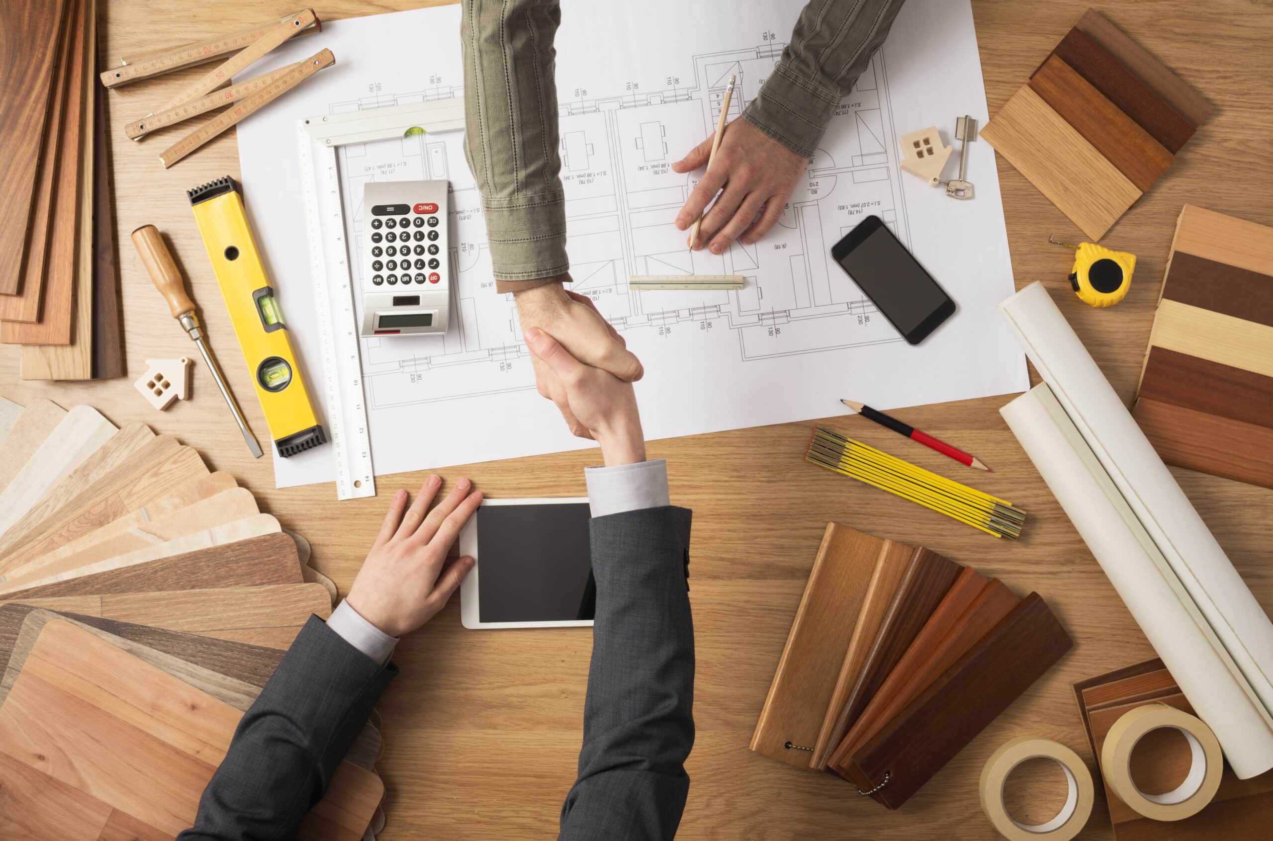 Architect and customer businessman Nearshoring shaking hands top view, desktop with building project, tools and wood swatches on background