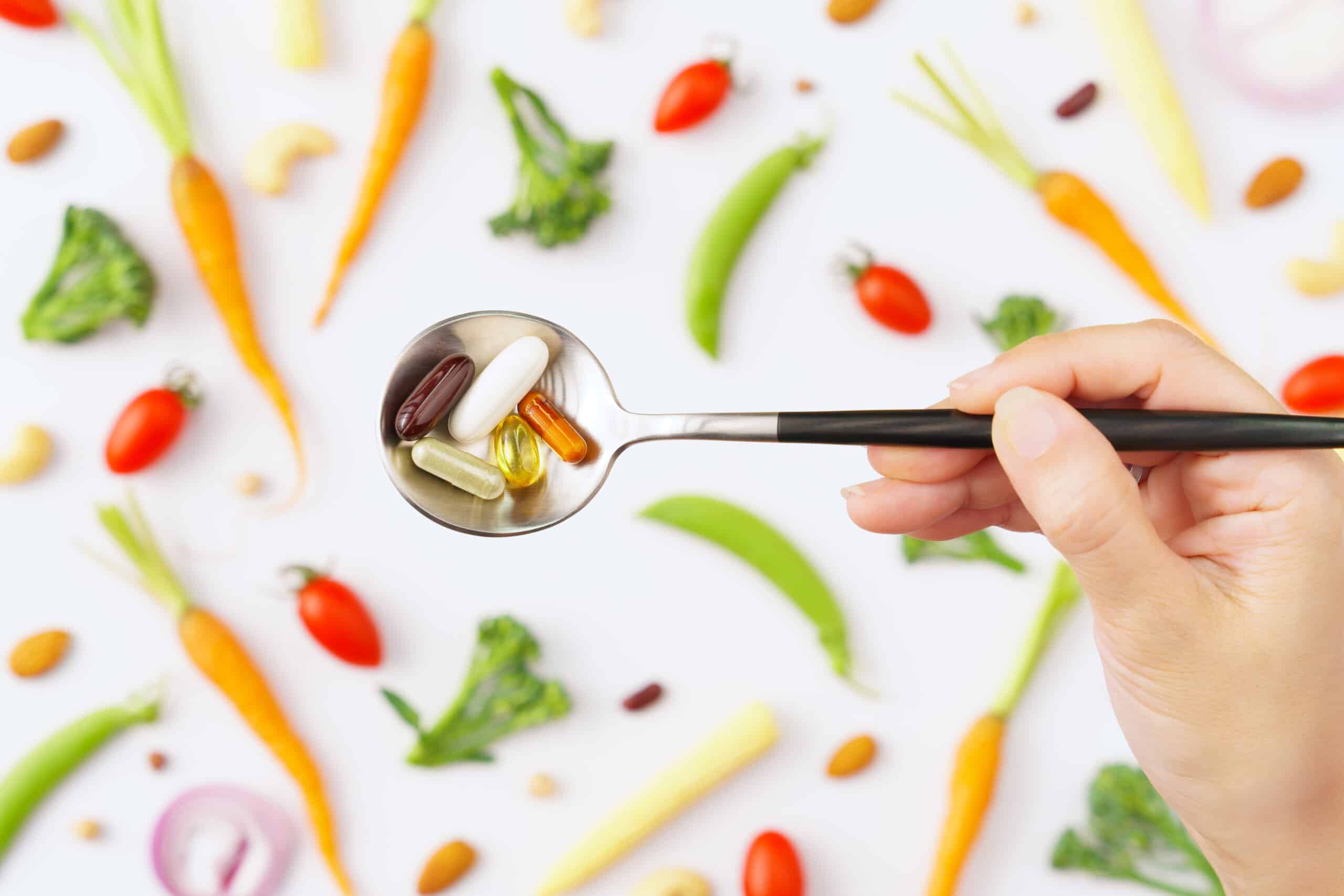 Balanced healthy vegan, plant based supplements concept. Top view close up of a spoon full of Vitamins and protein capsules with colorful phytonutrients vegetables as a background. Multivitamins, B12
