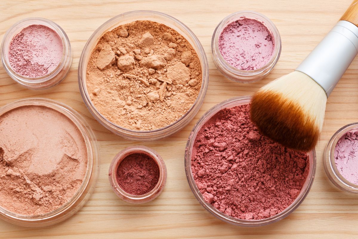 Bild, das einen Behälter mit losem Rougepigment neben verschiedenen anderen Make-up-Produkten zeigt, die alle ordentlich angeordnet sind, um ihre Farben und Texturen hervorzuheben, vor einem sauberen, einfachen Hintergrund.
