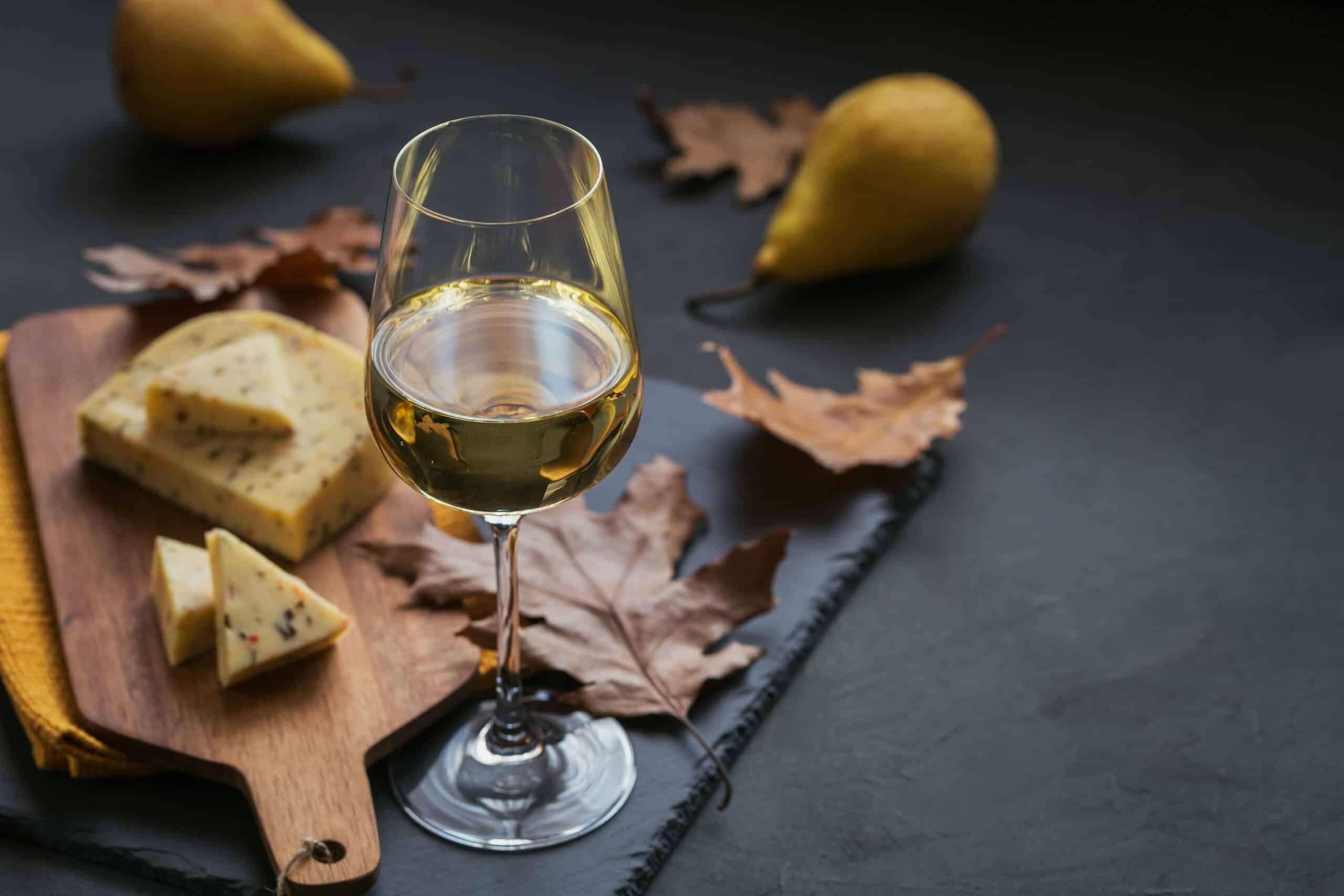 A glass of white wine was served with cheese in a cutting board on dark background. Autumn picnic with cheese, wine and dry leaves in rustic style.