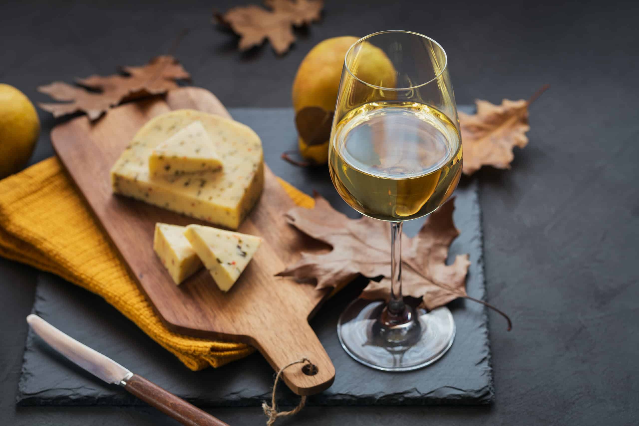 A glass of white wine was served with cheese in a cutting board on dark background. Autumn picnic with cheese, wine and dry leaves in rustic style.