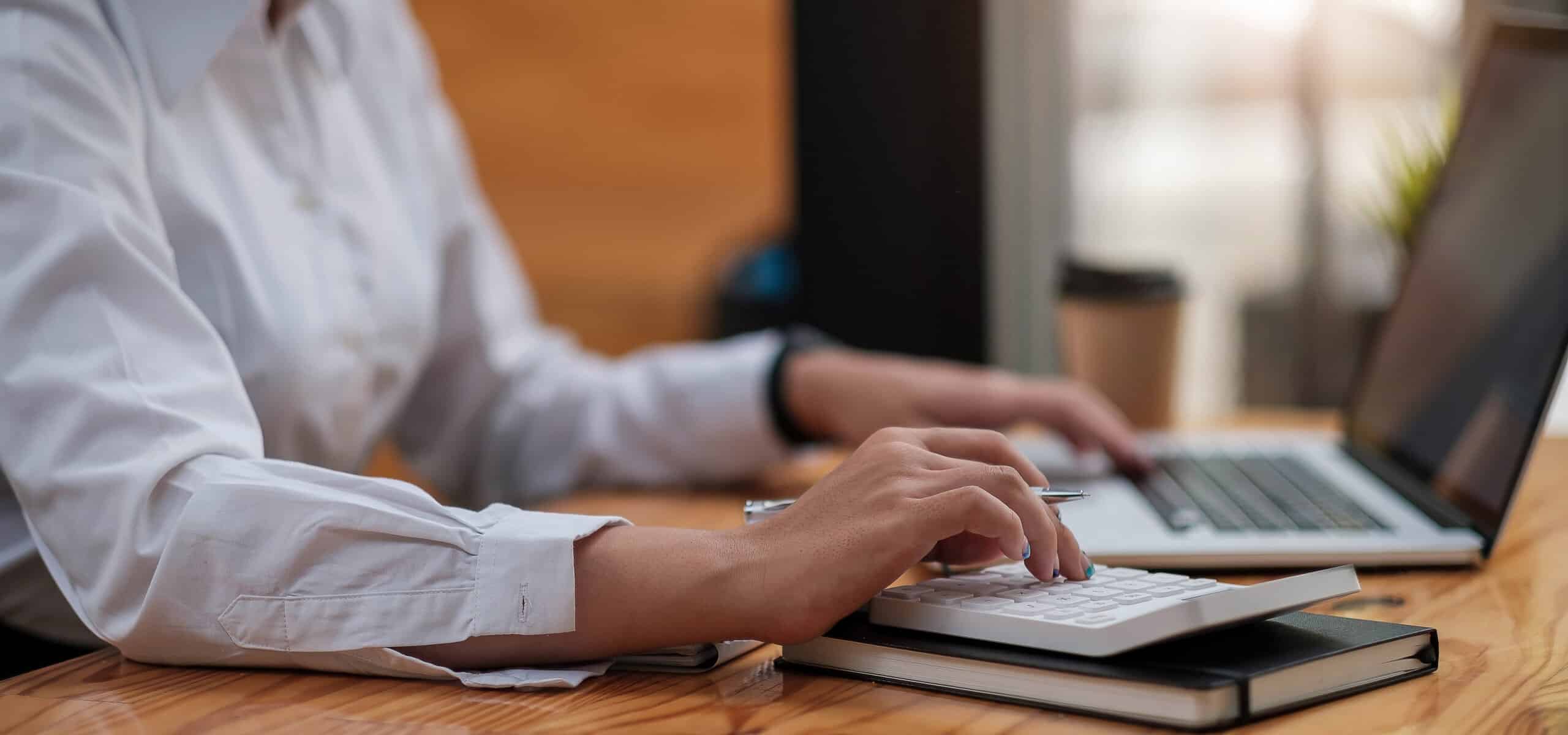 Eine Person, die an einem Schreibtisch mit einem Taschenrechner und einem Laptop arbeitet.