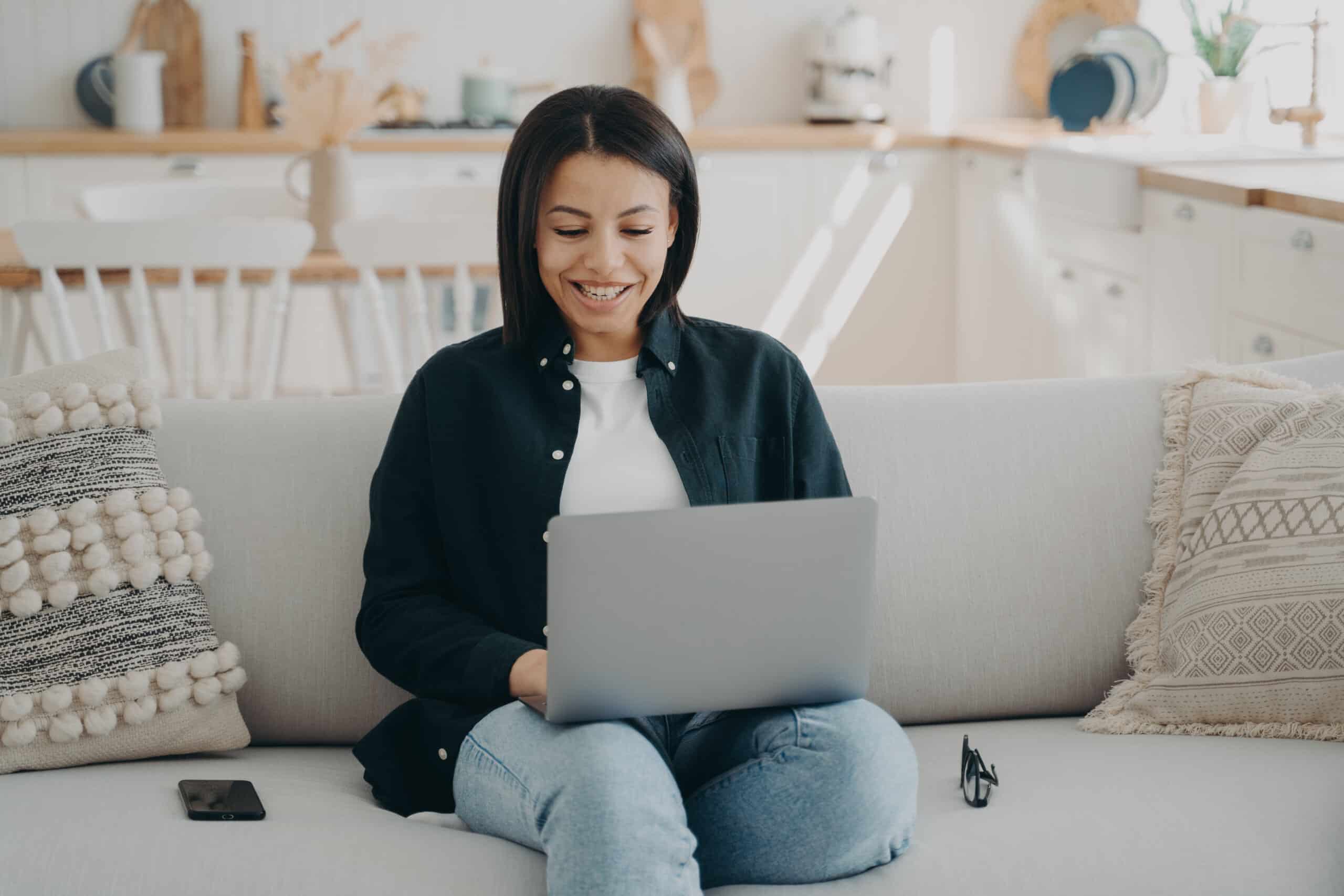 Smiling female shopping on the internet at laptop sitting on couch at home. Happy woman freelancer working on computer, chatting in social networks. Ecommerce, online store advertisement.
