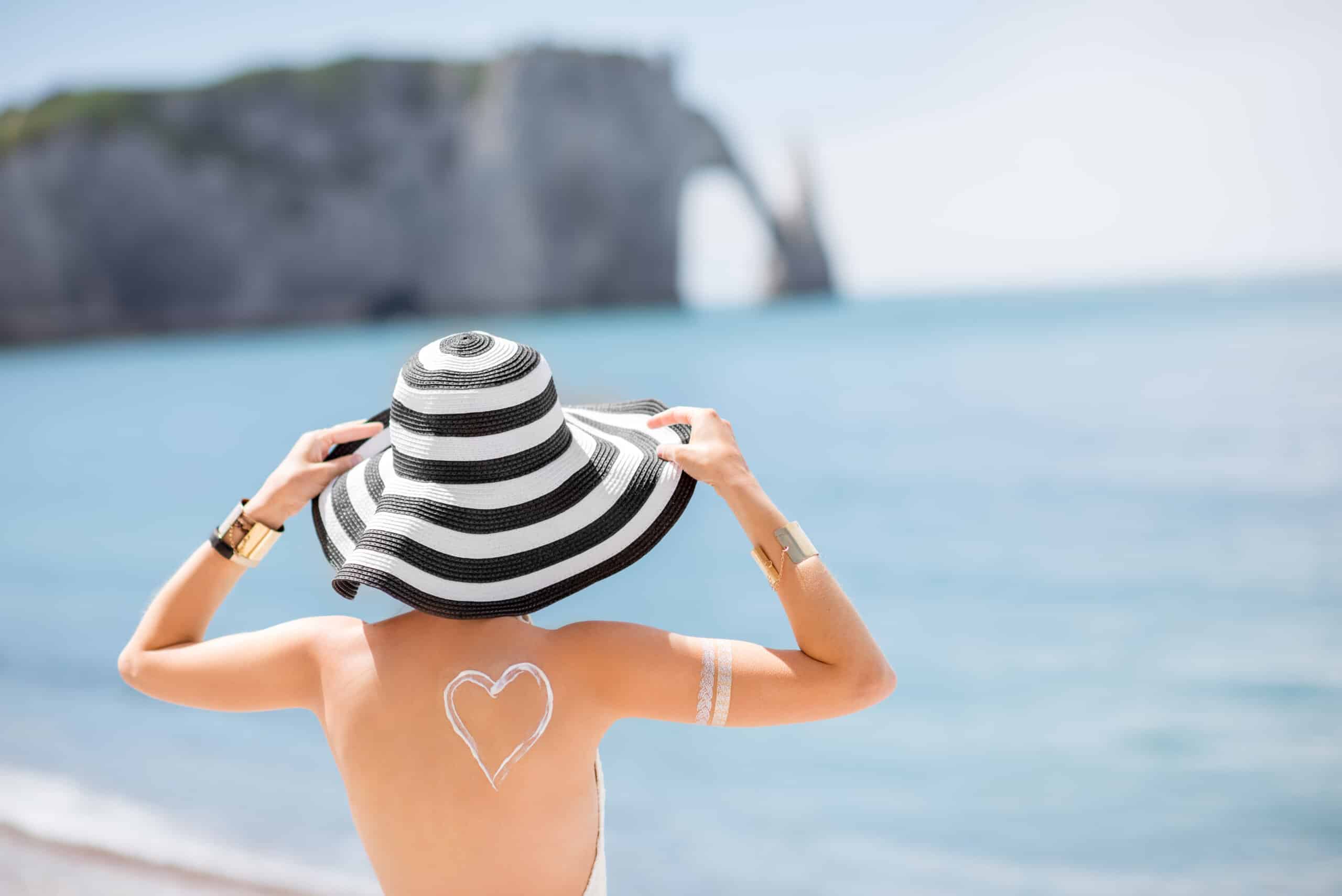 Rückansicht einer schönen Frau mit gestreiftem Hut und einem Herz aus Sonnencreme auf der Schulter, die sich am berühmten französischen Strand sonnt