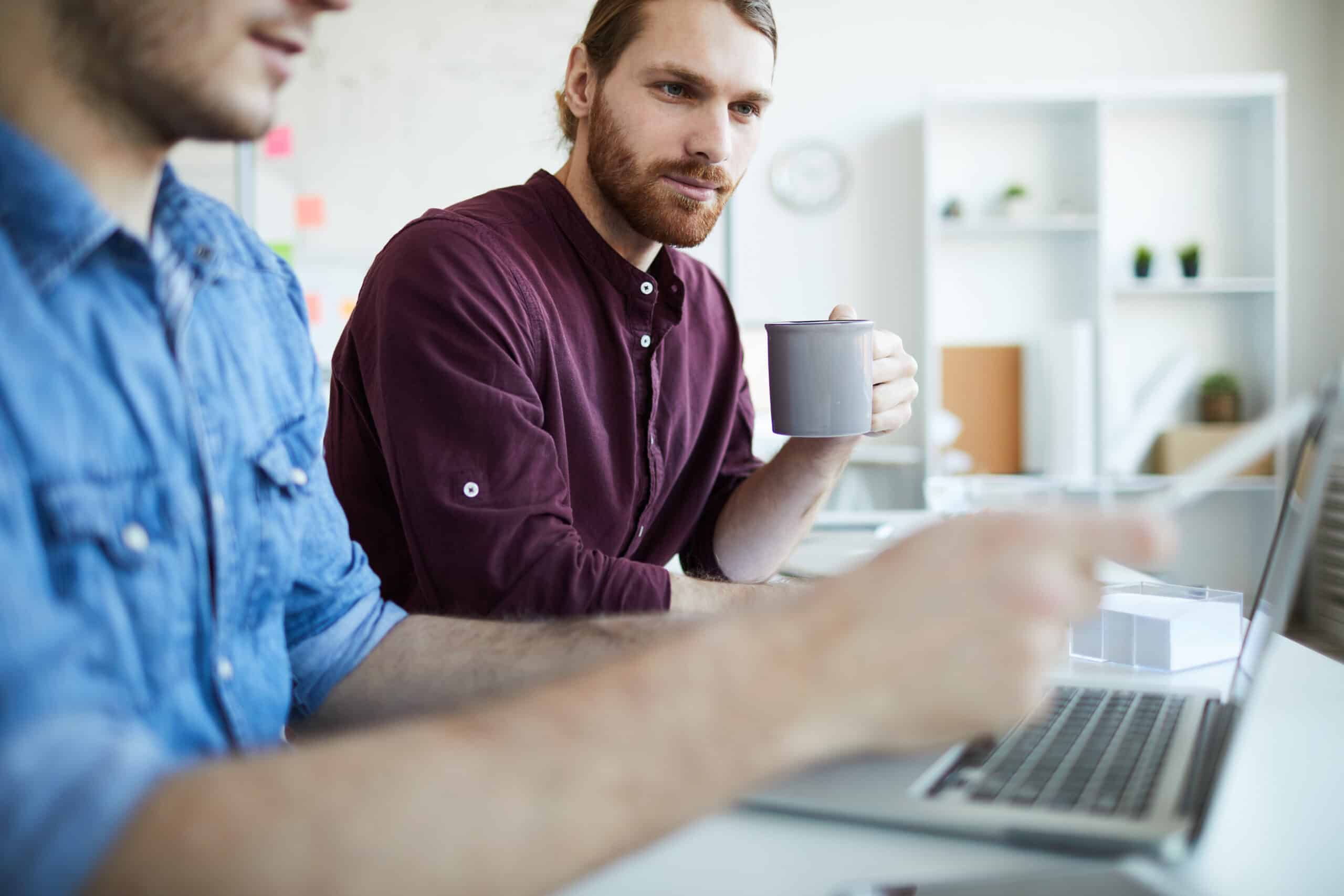 One of young casual webdesigners with mug looking at laptop display while his colleague making presentation of new website