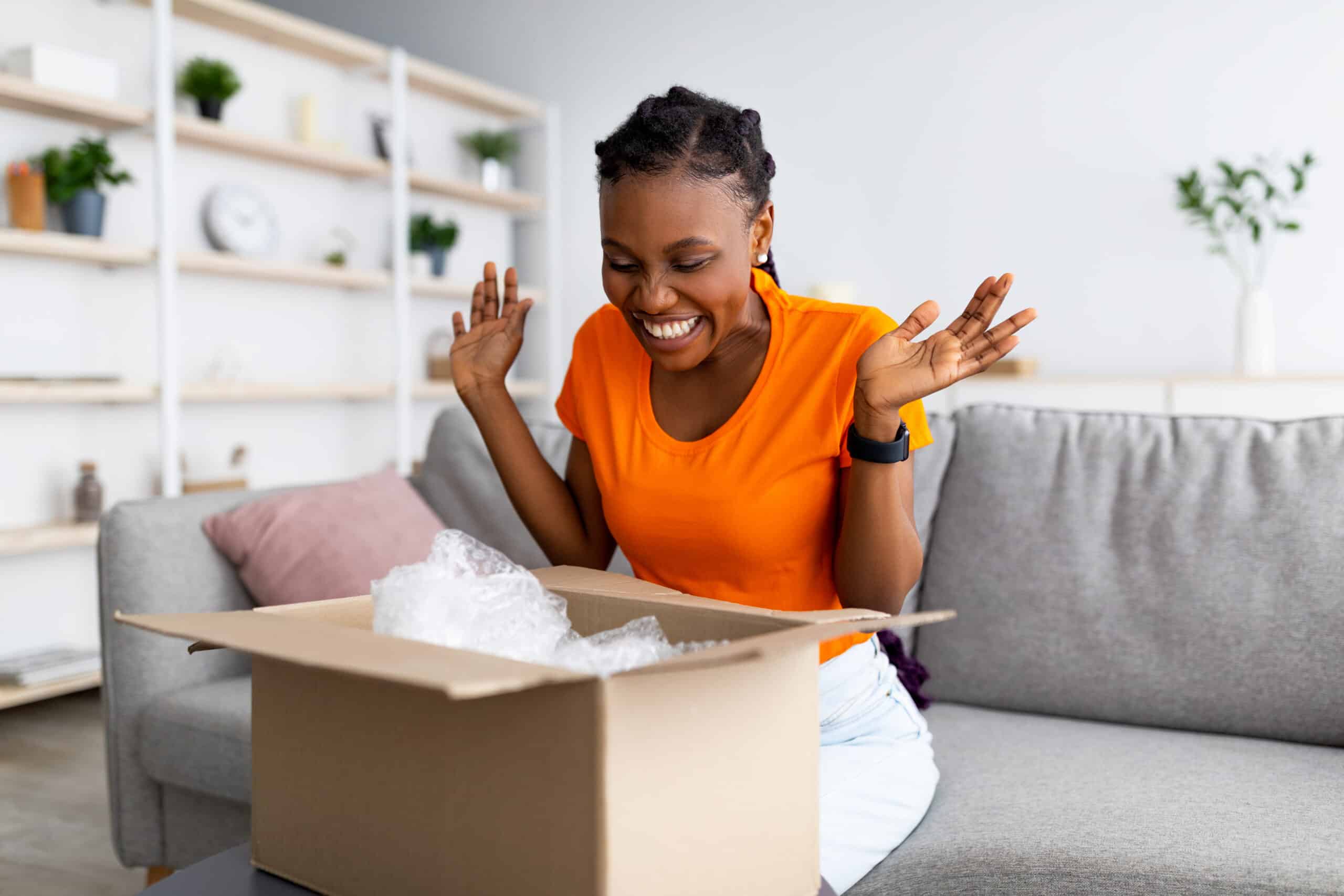 Overjoyed Afro woman unboxing carton parcel, emotional about successful shopping, satisfied with great purchase at home. Online store, reliable delivery service concept