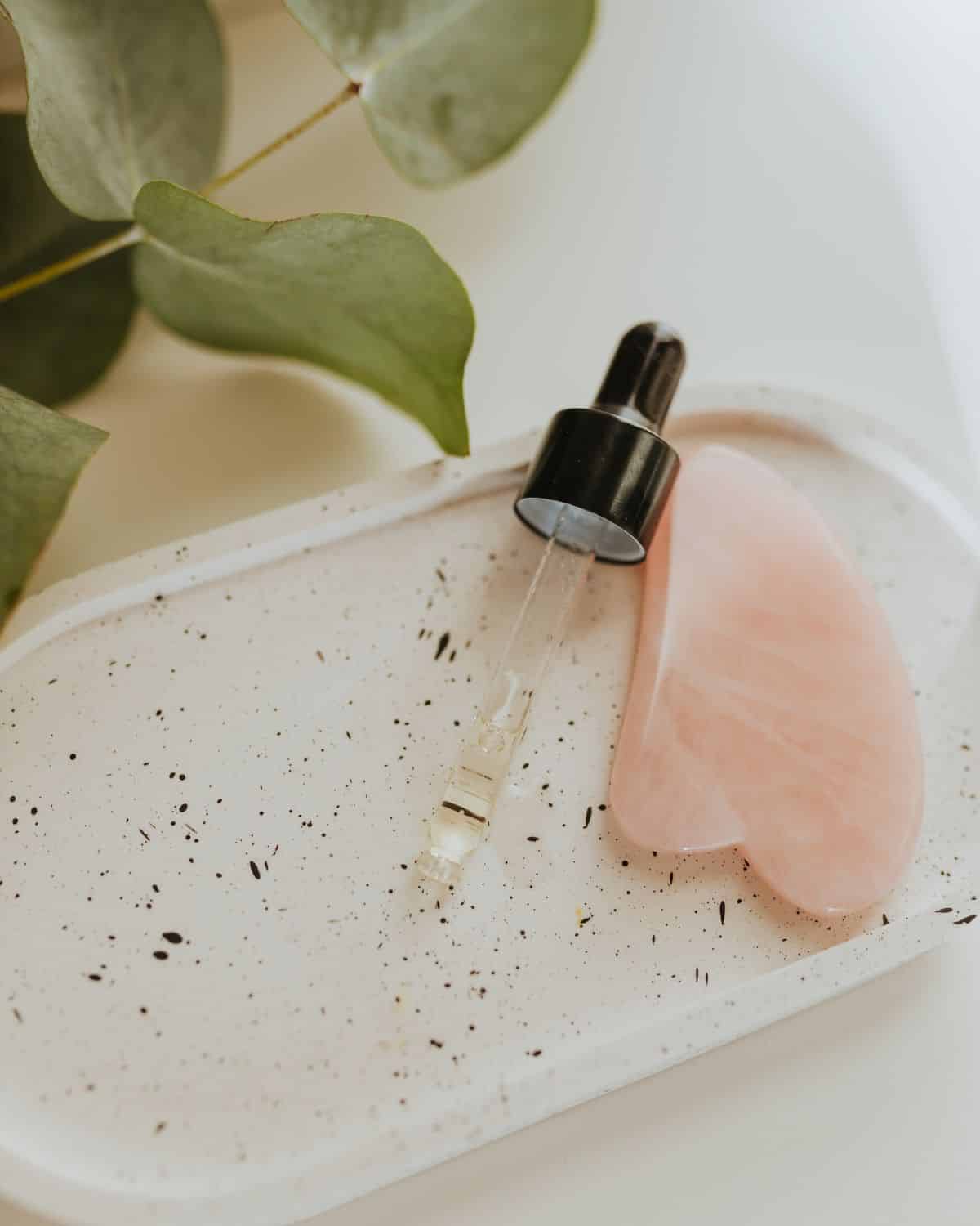 Nahaufnahme eines Gua-Sha-Steins neben einer Tropfflasche mit Gesichtsöl, die Wellness und Self-Care-Rituale im Hautpflegebereich illustriert.