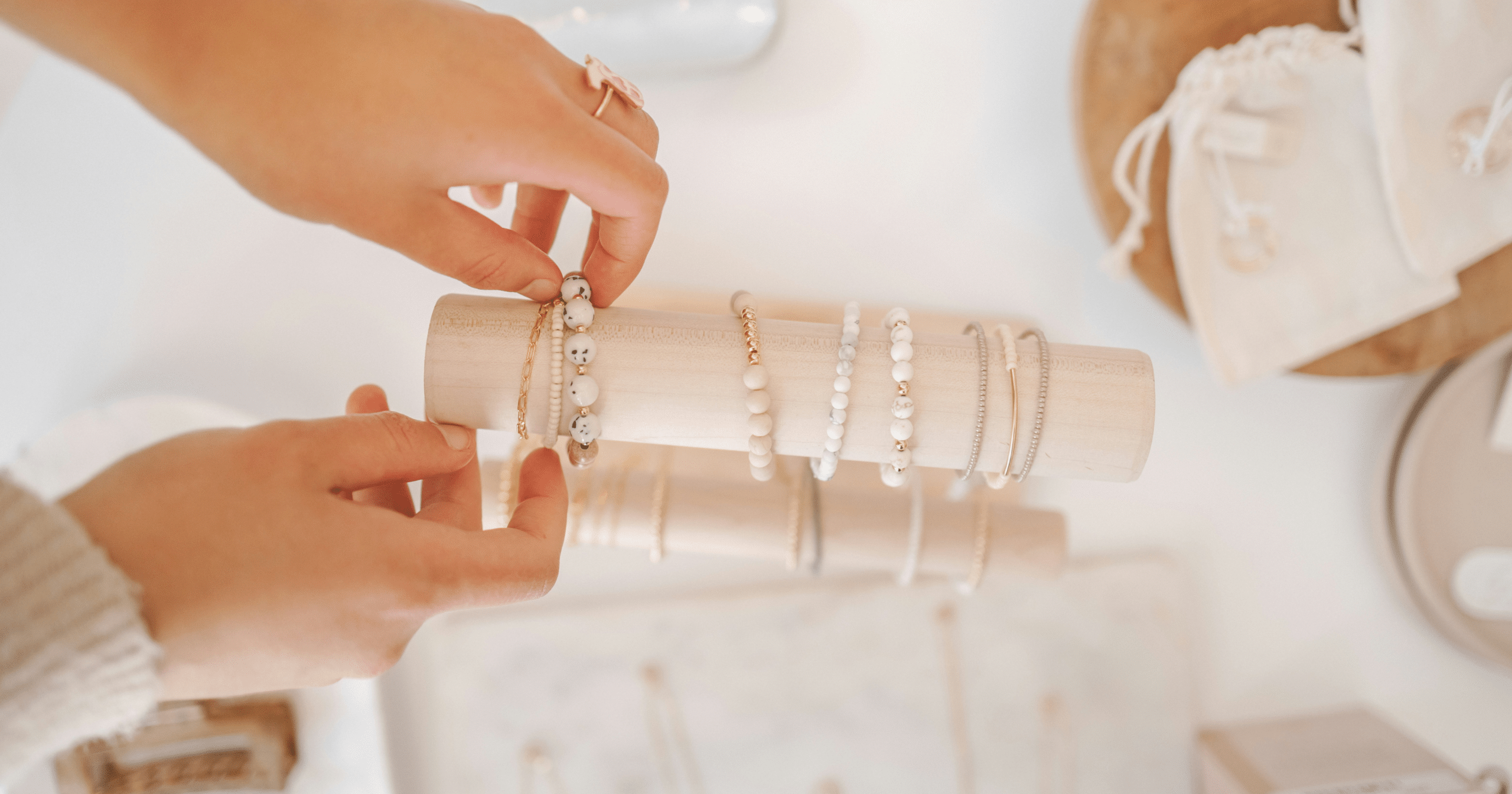 A person selecting jewelry from a display stand featuring elegant bracelets. Variety of white, gold, and beaded bracelets organized neatly, with additional jewelry items in soft pouches and boxes visible in the background. Luxurious and delicate aesthetic perfect for fashion and accessory enthusiasts.