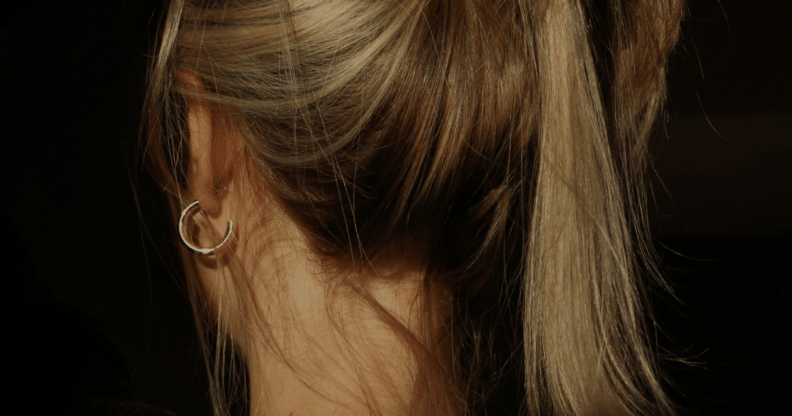 Woman with hair tied up in a casual ponytail, wearing a gold hoop earring. Hand resting gently on head, showcasing a natural and relaxed pose. 