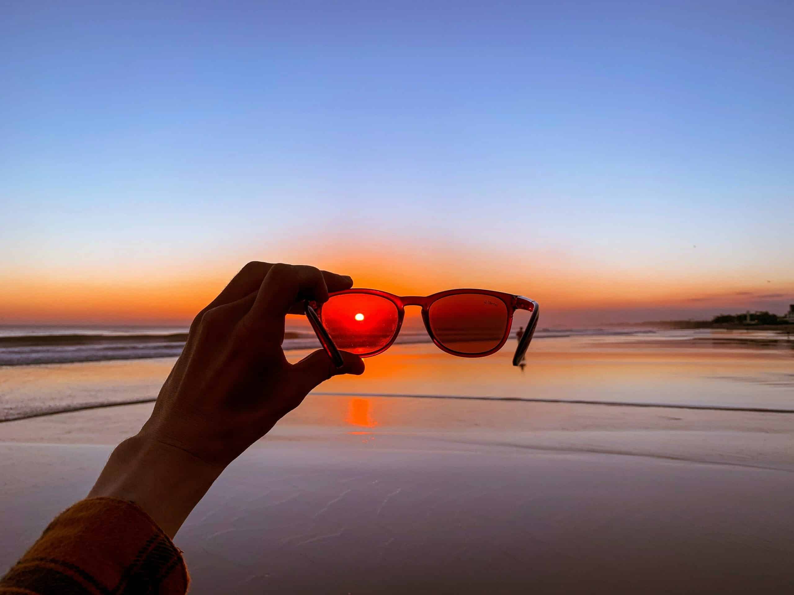 Eine Sonnenbrille, die von einer Person vor einer glamourösen Strandkulisse mit Sonnenuntergang gehalten wird. Die Sonne ist in der Linse der Brille platziert.