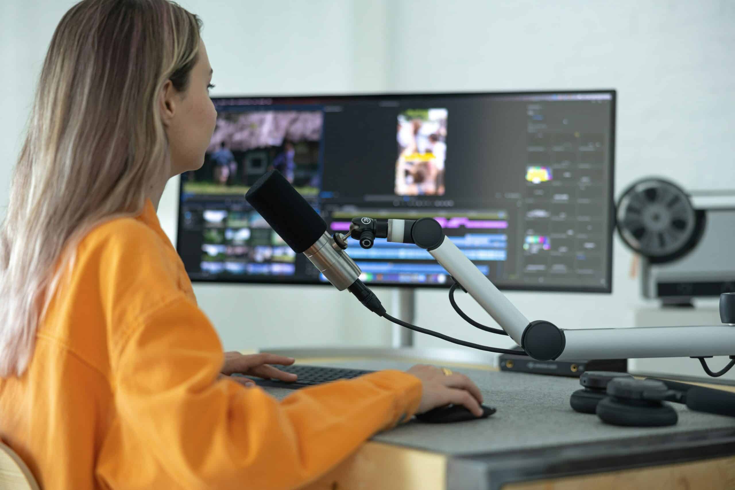A woman editing or filming part of a video, sitting at her desk speaking into a microphone with a computer in front of her.